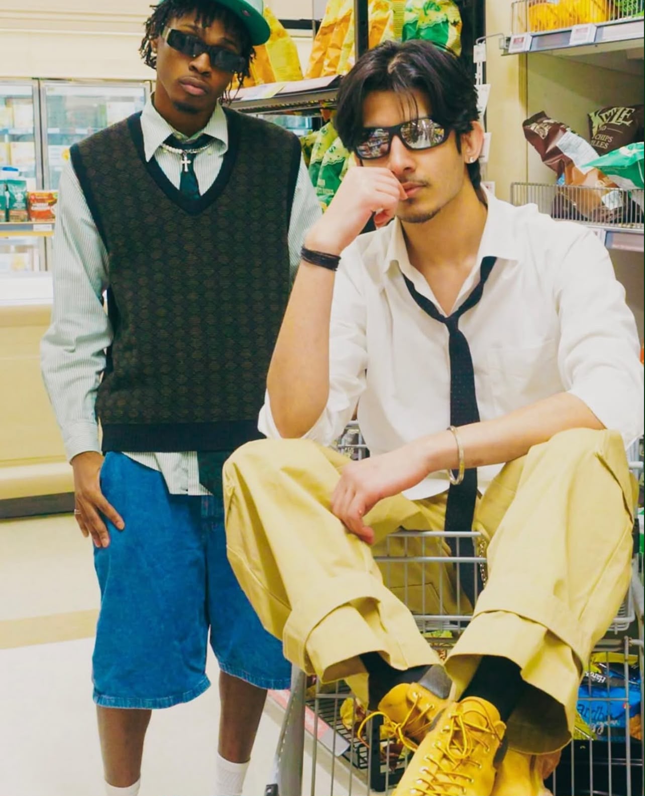 Two models in a shop, with the foreground model seated in a shopping cart wearing sunglasses and yellow trousers.