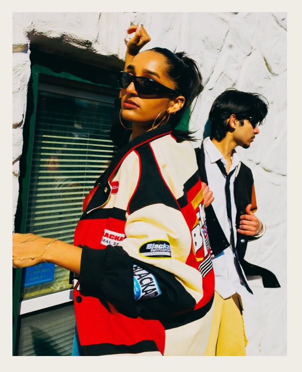 Two models wearing sunglasses and contrasting outfits by a white wall.